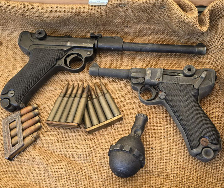Two vintage Luger pistols, bullets, and a grenade rest on a burlap surface in a historical weapon display.