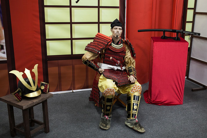 A lifelike samurai mannequin in red and gold armor sits in a traditional Japanese setting. 