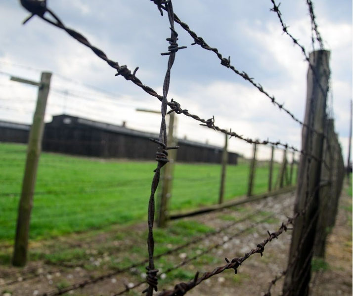 Close-up of rusty barbed wire fencing with wooden posts, enclosing a grassy area and distant dark buildings under a cloudy sky.