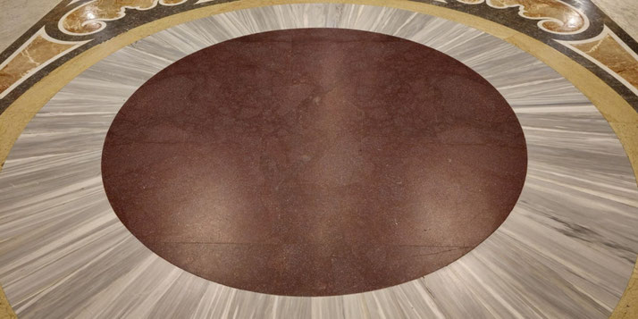 A circular floor design featuring a central reddish-brown marble disk surrounded by radiating gray marble segments, bordered with ornate detailing.