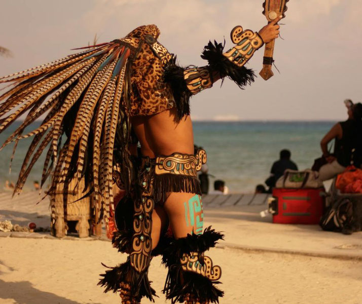 A performer dressed as an Aztec Jaguar Warrior stands on a tropical beach, wearing a jaguar headdress, feathered adornments, and ornate armor while holding a macuahuitl club in a dramatic pose.