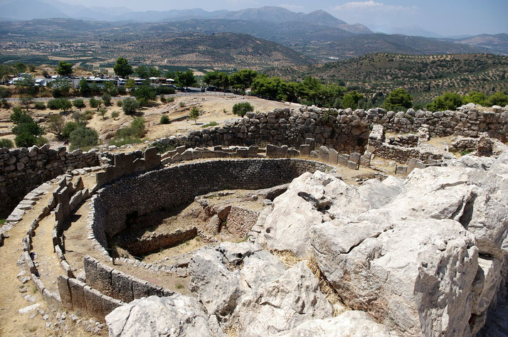 Circular stone grave enclosure at the archaeological site of Mycenae, surrounded by rocky terrain and distant hills.