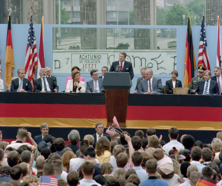 Ronald Reagan delivers a speech near the Berlin Wall before a crowd, flanked by U.S. and German officials and flags.