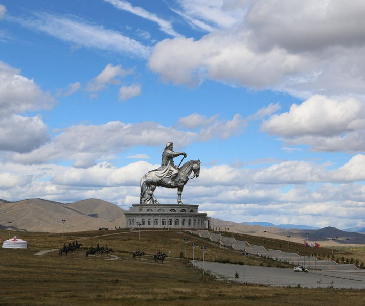 Long distance photo of a statue of Genghis Khan, a massive stainless-steel monument, stands against a vast, open landscape with rolling hills, yurts, and horsemen under a bright blue sky with clouds.