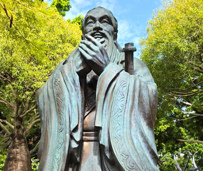 A bronze statue of a smiling Confucius in traditional robes, clasping his hands together in a gesture of wisdom and respect, set against a backdrop of lush green trees and a bright blue sky.
