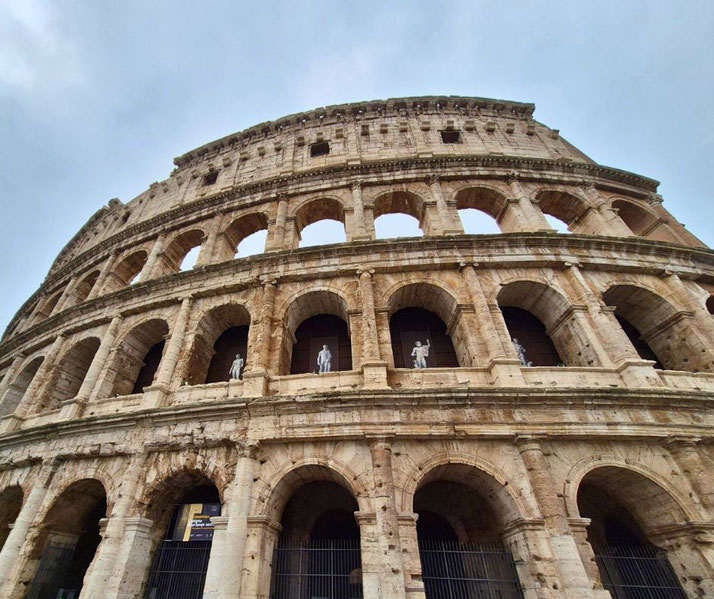 The exterior of the Colosseum from a low angle, showcasing its iconic arches and weathered stone. Statues are visible in some openings.