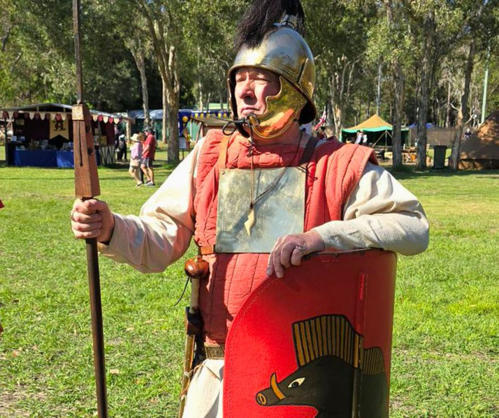 Man in Roman soldier costume holds a spear and red shield with boar emblem, standing in a grassy area at a historical reenactment event.