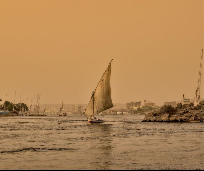 A sailboat moves through calm waters at sunset with city buildings and other boats in the background under an orange sky.