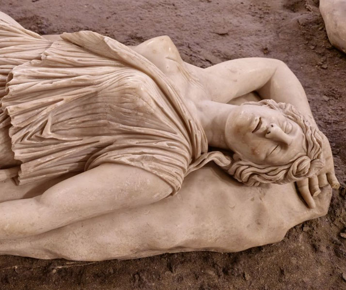 Marble statue of a reclining woman with closed eyes, draped in flowing robes, lying on a stone cushion with a peaceful, serene expression.