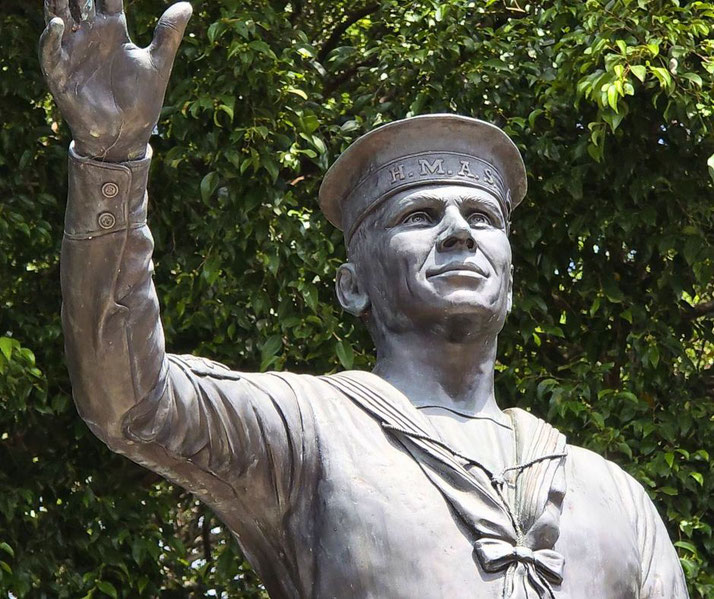 A bronze statue of a smiling sailor in HMAS uniform raises one hand in a gesture of farewell or greeting.