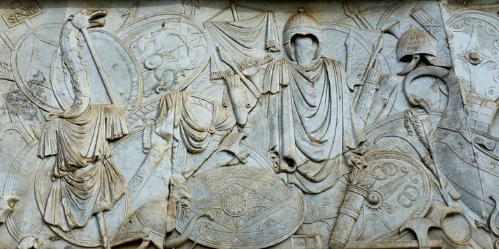 Ancient Roman marble relief showing armor, shields, helmets, standards, and draped garments, symbolizing military strength and victory.