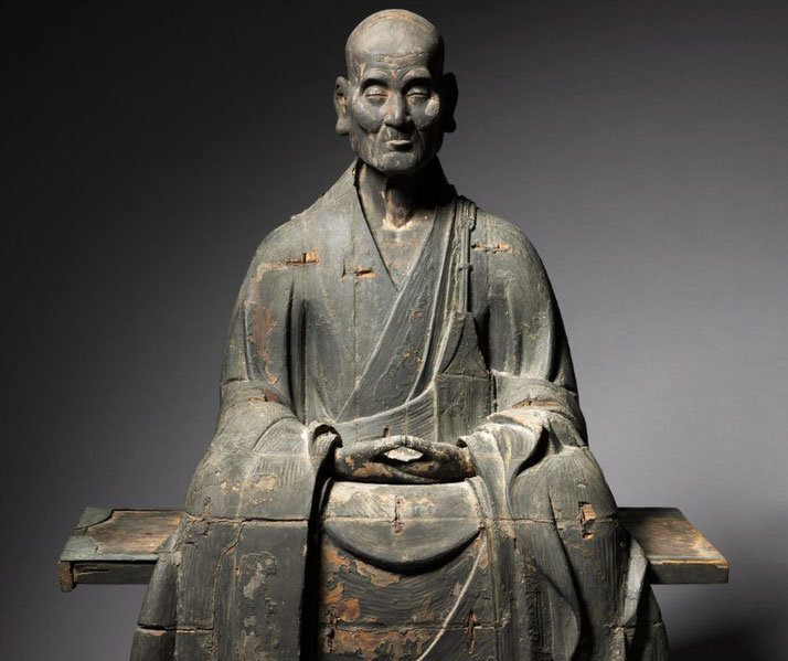 A weathered wooden statue of a seated Buddhist monk, wearing traditional robes. The figure has a calm, meditative expression with hands gently resting in his lap.