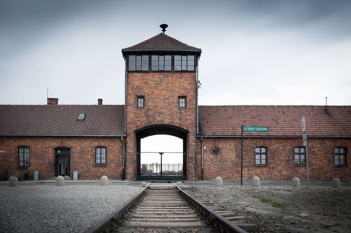 Railway tracks lead directly into the brick entrance gate and watchtower of a historical concentration camp under a cloudy sky.