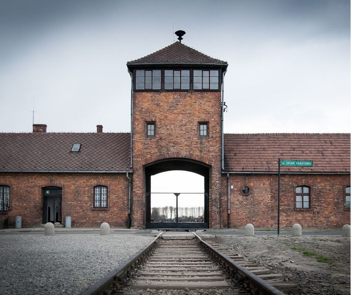 Railway tracks lead directly into the brick entrance gate and watchtower of a historical concentration camp under a cloudy sky.