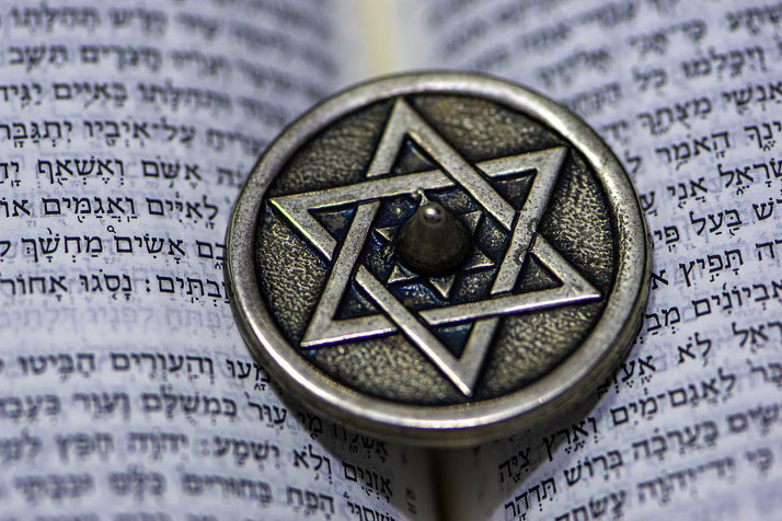 A metallic dreidel with a Star of David design rests on an open Hebrew text, with focused detail on both elements.