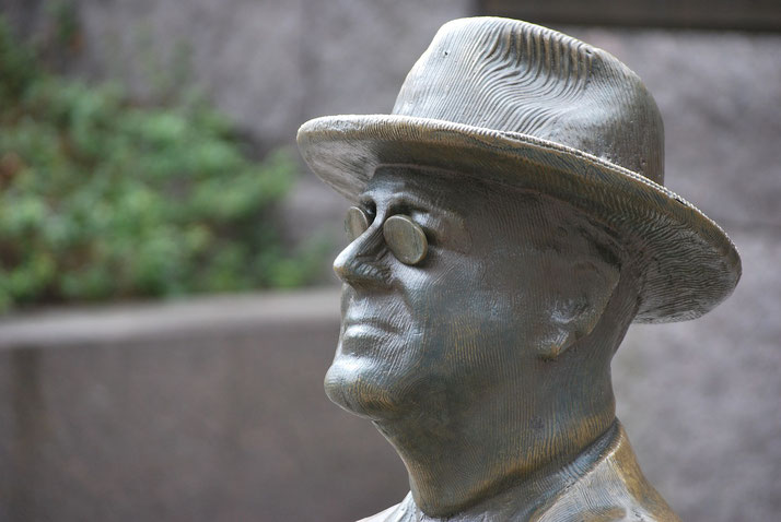 A bronze statue of a man wearing round glasses and a hat gazes upward with a composed expression, outdoors.