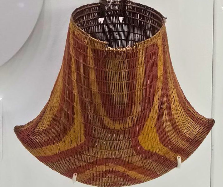 A beautifully crafted woven basket with flared edges, featuring intricate patterns in earthy red, yellow, and brown tones, displayed in a well-lit museum exhibit.