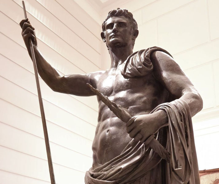Bronze statue of a muscular man in a heroic pose, holding a spear and draped in a cloak, symbolizing strength and leadership.