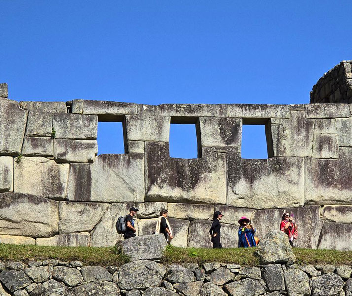 A section of Machu Picchu’s ancient stone walls with rectangular windows, set against a clear blue sky. Visitors, including some in traditional Andean clothing, walk along the historic site.