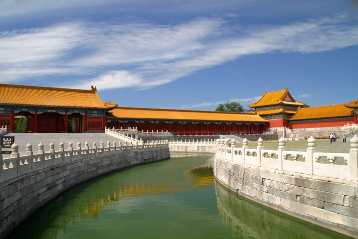 A scenic view of the Forbidden City in Beijing, featuring golden-roofed buildings, red walls, a curved waterway, and a white stone bridge under a bright blue sky.