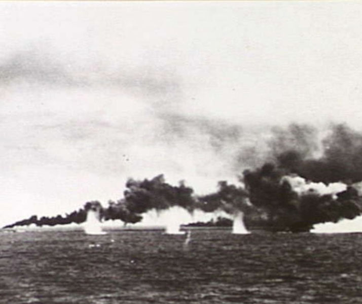 A warship is under attack at sea, engulfed in thick smoke and explosions as water splashes from nearby shell impacts.