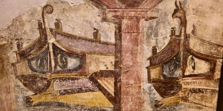 Ancient fresco depicting two Roman warships with decorated prows, painted eyes, and figureheads, docked beside a stone column.