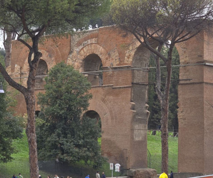 Ancient Roman brick aqueduct arches partially preserved, surrounded by trees and greenery in a public area.