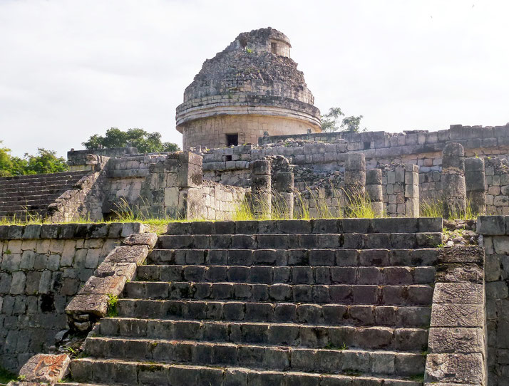 "El Caracol," an ancient Mayan observatory in Chichén Itzá, Mexico. The round stone structure, partially ruined, sits atop a platform with staircases, surrounded by overgrown vegetation.