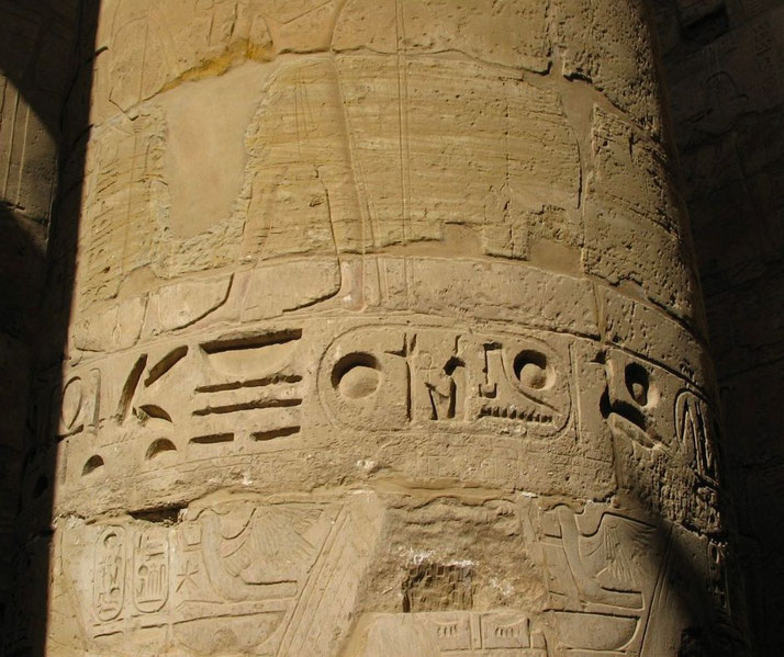Close-up of a weathered stone column engraved with detailed ancient Egyptian hieroglyphs and faded relief figures.