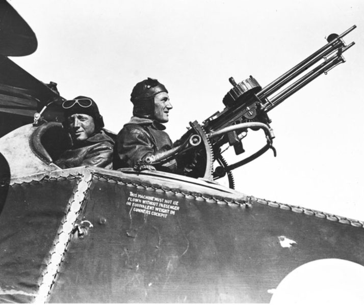 Two pilots in leather gear sit in a biplane; the rear gunner is manning a twin-barrel machine gun pointed skyward.