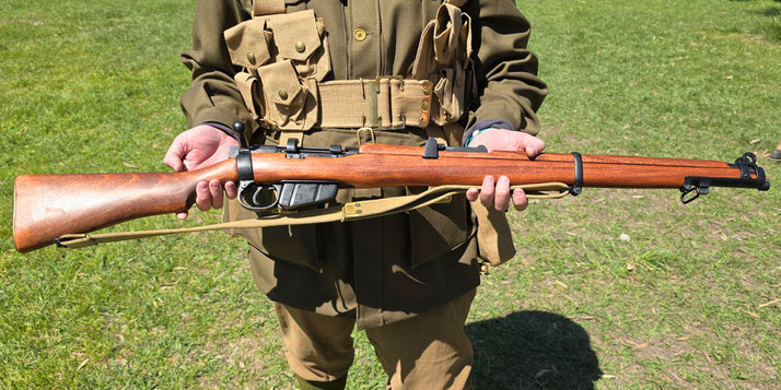 A person in vintage military uniform holds a bolt-action Lee-Enfield rifle with a wooden stock and mounted sling, standing on grass.