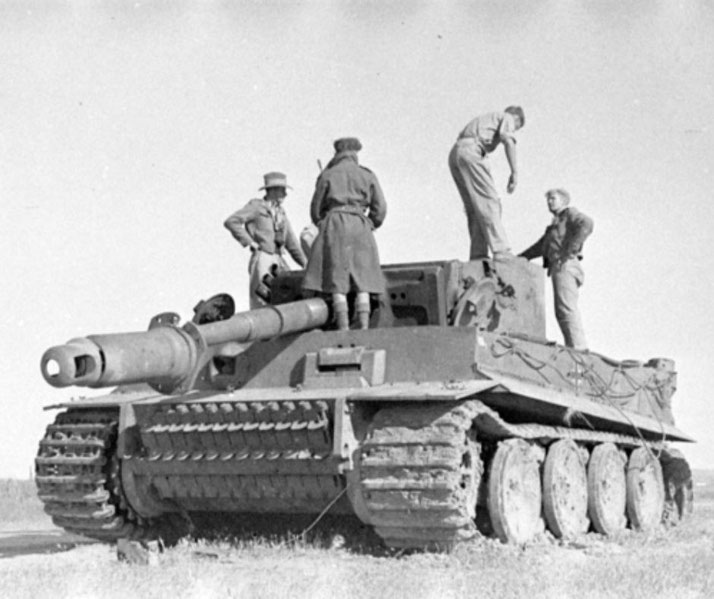 Soldiers inspect a large tank with wide tracks and a heavy gun, standing on and around it in an open field.