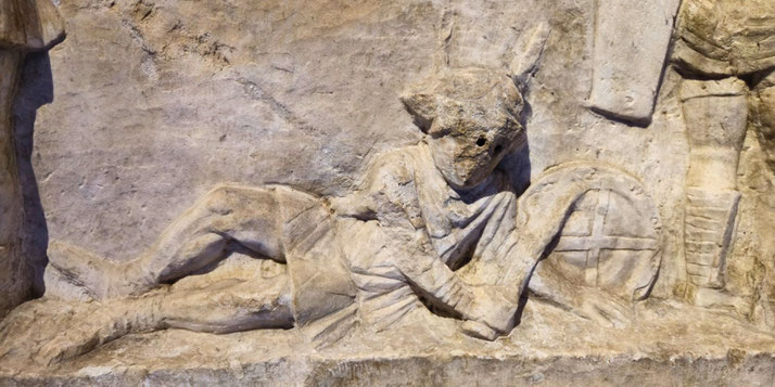 Close-up of a relief showing a fallen gladiator in a helmet and short tunic, lying on his side beside a round shield with a cross design.