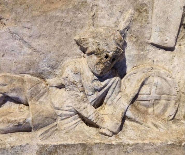 Close-up of a relief showing a fallen gladiator in a helmet and short tunic, lying on his side beside a round shield with a cross design.