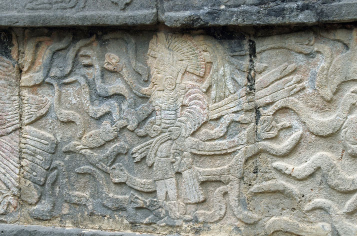 An ancient Mayan stone carving featuring a warrior or deity adorned with an elaborate headdress and armor.