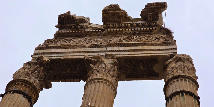 Ruins of an ancient Roman temple with three Corinthian columns and an intricately carved entablature, partially broken but rich in floral and geometric detail.