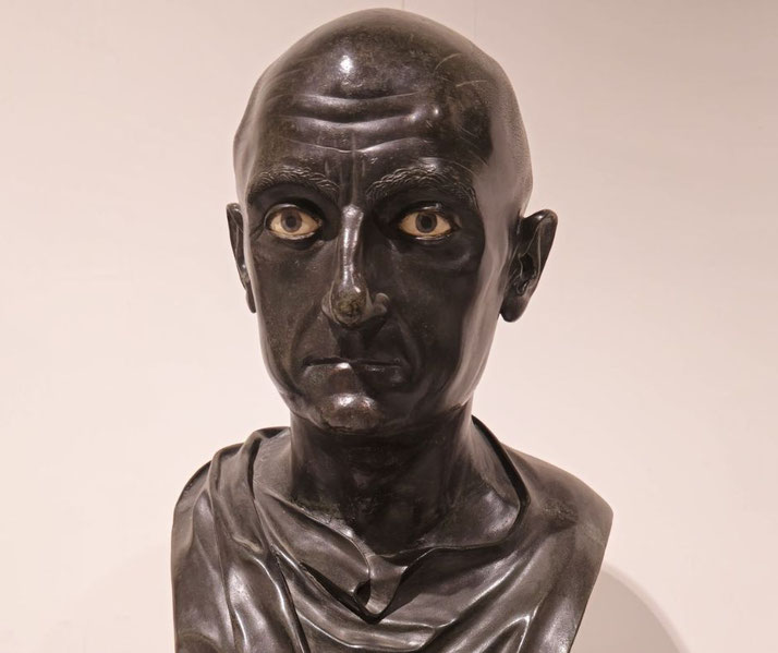 Bronze Roman bust of a stern, bald man with inlaid eyes, wearing a toga, showcasing intense realism and detailed facial features.