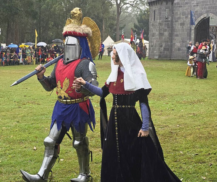A medieval reenactment with a knight in armor and a red tunic featuring a lion crest, accompanied by a woman in period attire. They walk hand-in-hand near a stone castle and festival crowd.