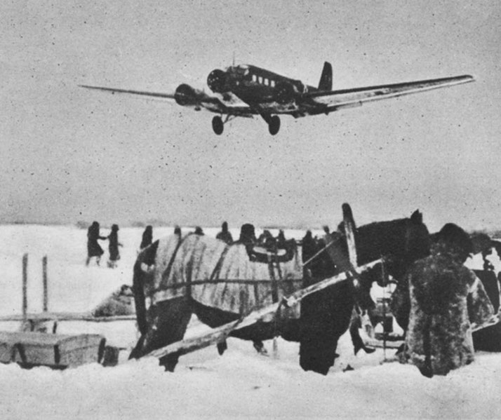 A low-flying aircraft approaches a snowy field where people and a horse-drawn sled are gathered below.