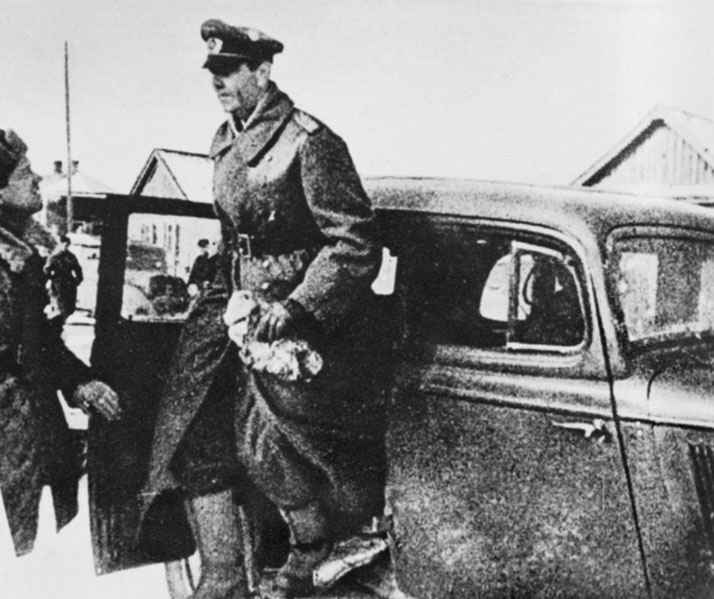 A military officer in uniform steps out of a car while another person stands nearby, both dressed for cold weather.