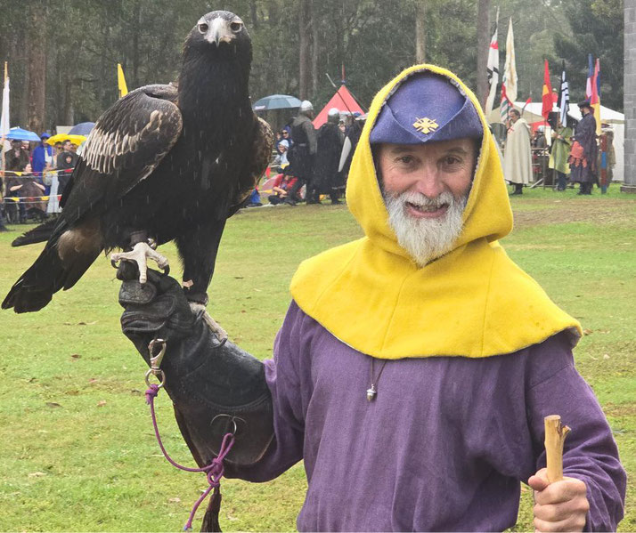 Man in medieval attire smiles while holding a large bird of prey on a gloved hand at a historical reenactment event.