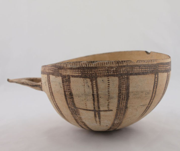 An ancient ceramic bowl from the Late Bronze Age, featuring a geometric pattern in dark pigment. The vessel has a handle and a small chip on the rim, indicating age and use.