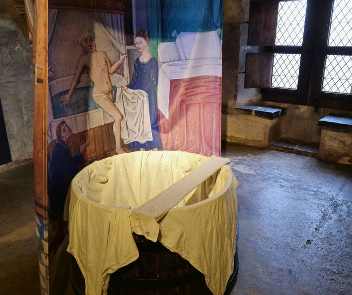 A medieval-style wooden bath with cloth lining sits in a historic stone room. A tapestry depicting a bathing scene adds historical context, evoking medieval hygiene and daily life in a castle setting.