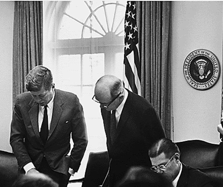 President John F. Kennedy and advisors are seen in the Oval Office during a tense meeting, with the presidential seal and U.S. flag visible in the background.
