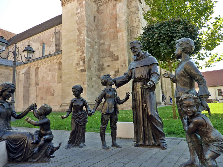 A bronze statue group of a monk interacting with children and a mother with a baby, set against a historic stone church.