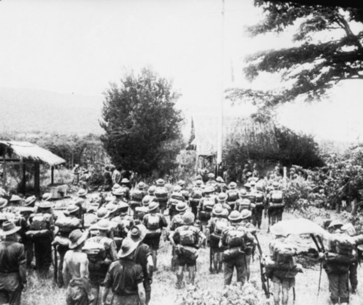 Black-and-white photo showing a large group of soldiers with backpacks and hats gathered in a rural setting near thatched huts.