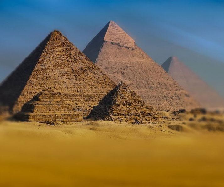The Pyramids of Giza, with a clear blue sky and golden desert sands. The foreground features smaller pyramids, while the iconic Great Pyramid dominates the background.