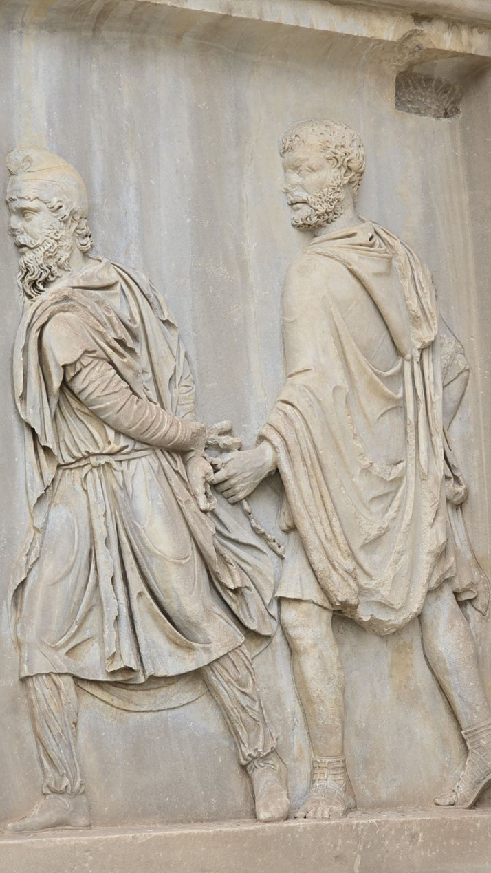 Stone relief sculpture of a bearded man being led in chains by another figure, both dressed in Roman-style garments.