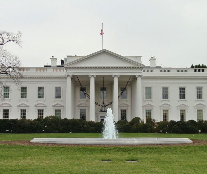 The White House stands with a U.S. flag atop, fronted by a fountain and lawn, under an overcast sky.
