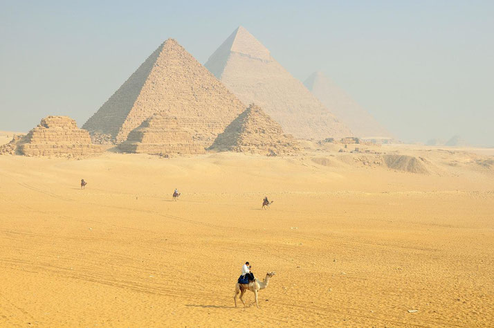 Camels and riders cross the sandy foreground with the pyramids of Giza rising behind them under a hazy sky.